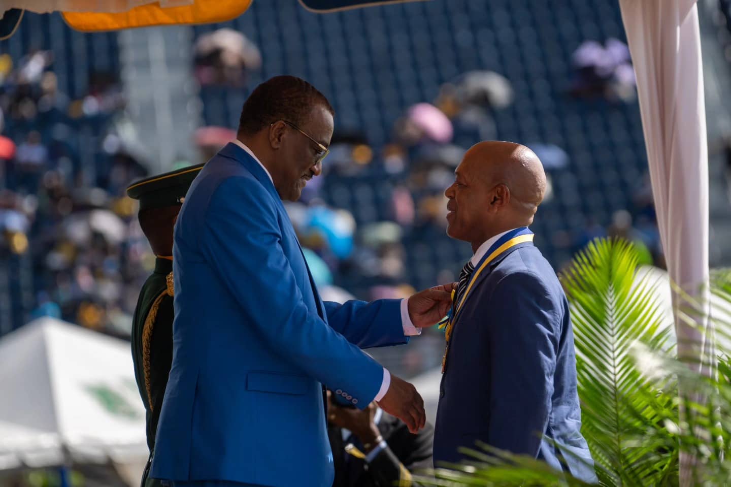 Two men in suits shaking hands at an outdoor ceremony with spectators in the background.