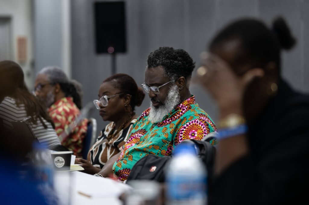 Conference attendees seated at a meeting table, including a man with a gray beard in a colorful patterned shirt, listening.