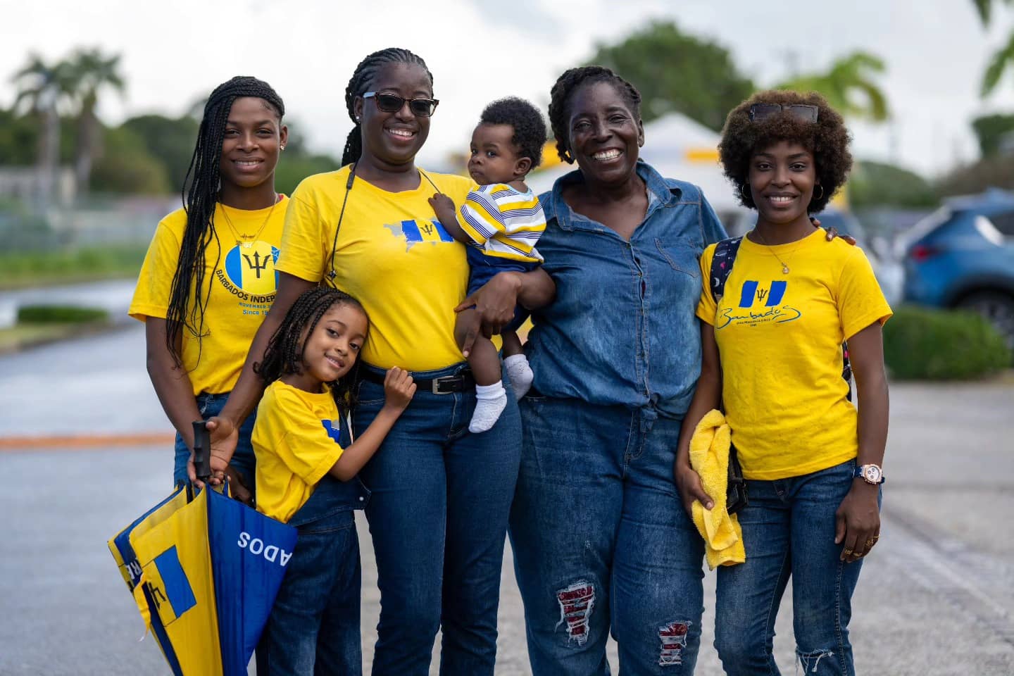Women and children wearing yellow shirts standing outdoors with a Barbados flag.
