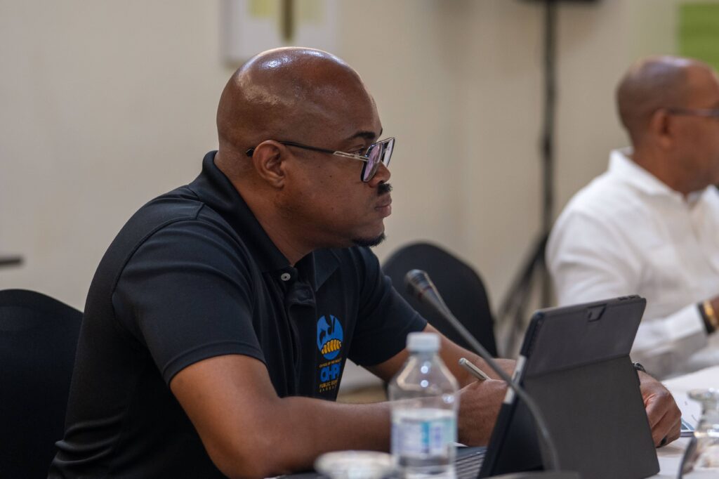 Man in a black polo shirt speaking and gesturing at a conference table with a laptop and microphones.