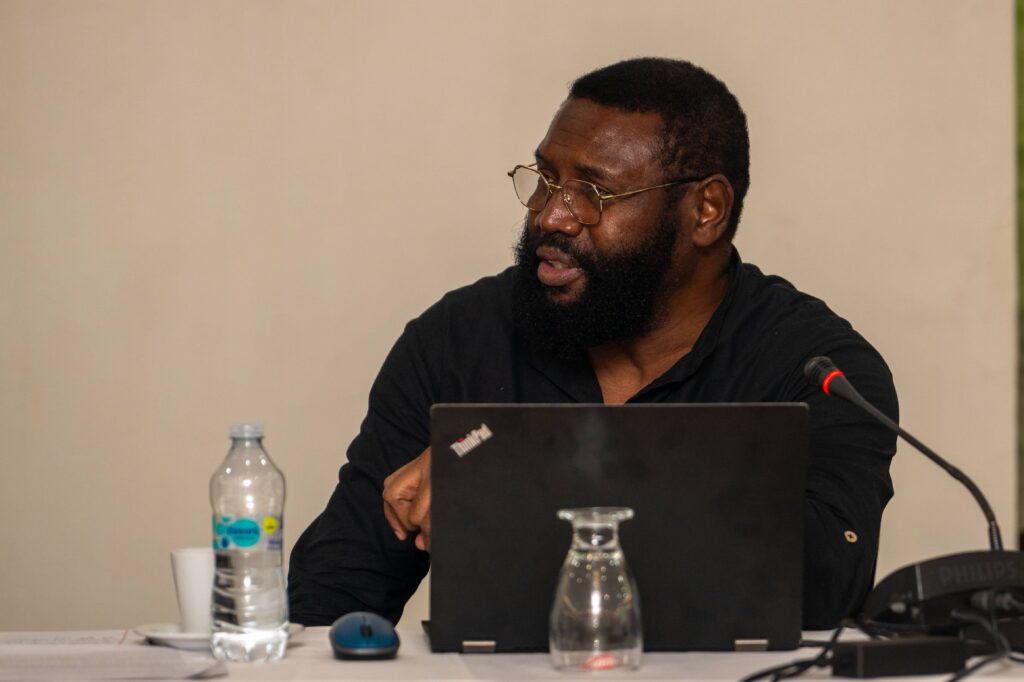 Bearded man wearing glasses speaking at a conference table using a laptop, with a microphone and water bottle on the table.