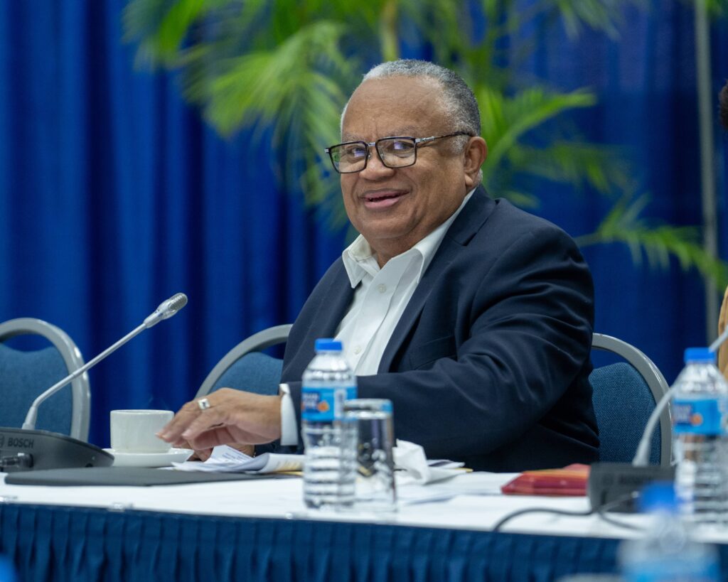 Smiling man seated at a conference table during a meeting with a microphone and water bottles.