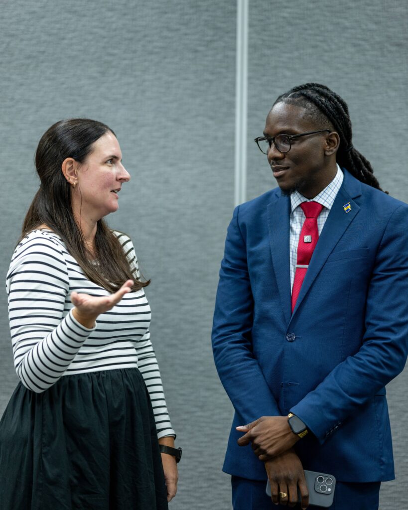 Two people standing and talking; a woman gesturing with her hand and a man in a blue suit with a red tie listening.